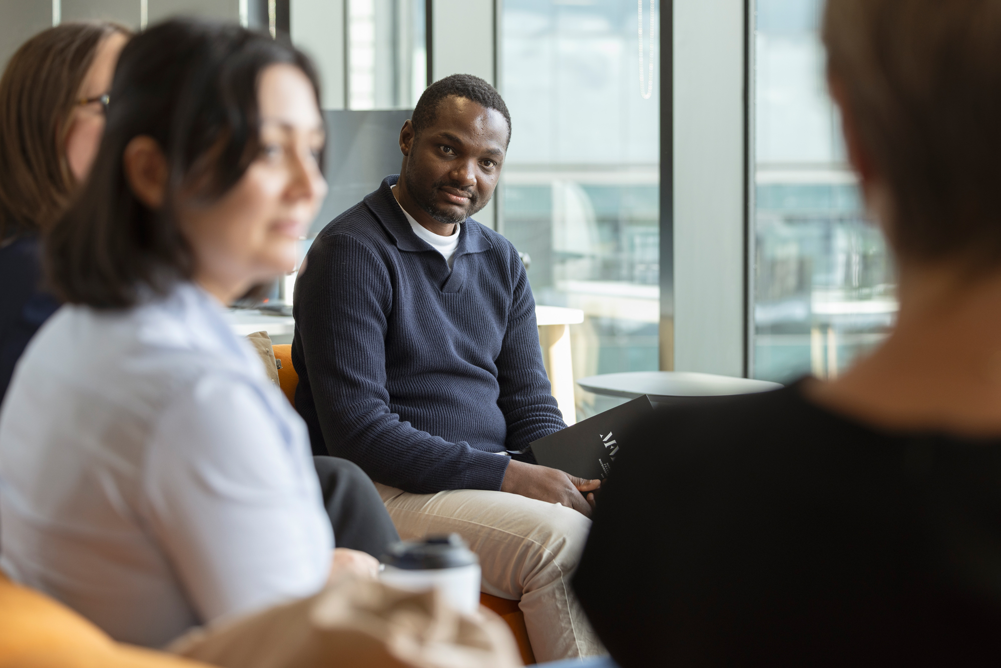 A group of professionals seated in a circle in a well-lit office, engaged in a discussion. One person holds a notebook and looks toward the camera, while others focus on the conversation.