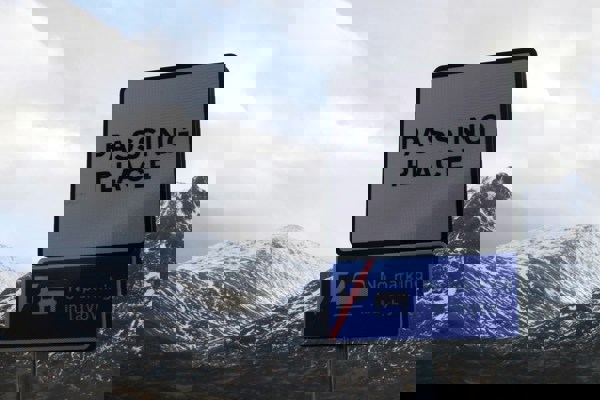 Roadside sign reading “PASSING PLACE” above another sign stating “No parking in layby,” set against a backdrop of snow-capped mountains and cloudy sky in a rural Scottish landscape.