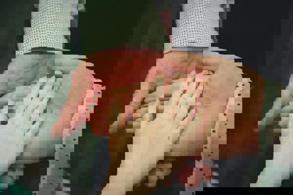 Two adult hands held palm up with a child's hand resting gently on one of them. The adults are wearing patterned long-sleeved shirts—one with small black dots and the other with a grid-like design.